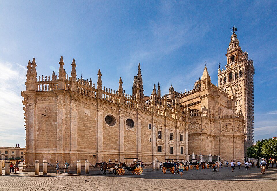 The cathedral from the square, with the Giralda rising at its far corner and horse carriages waiting below.